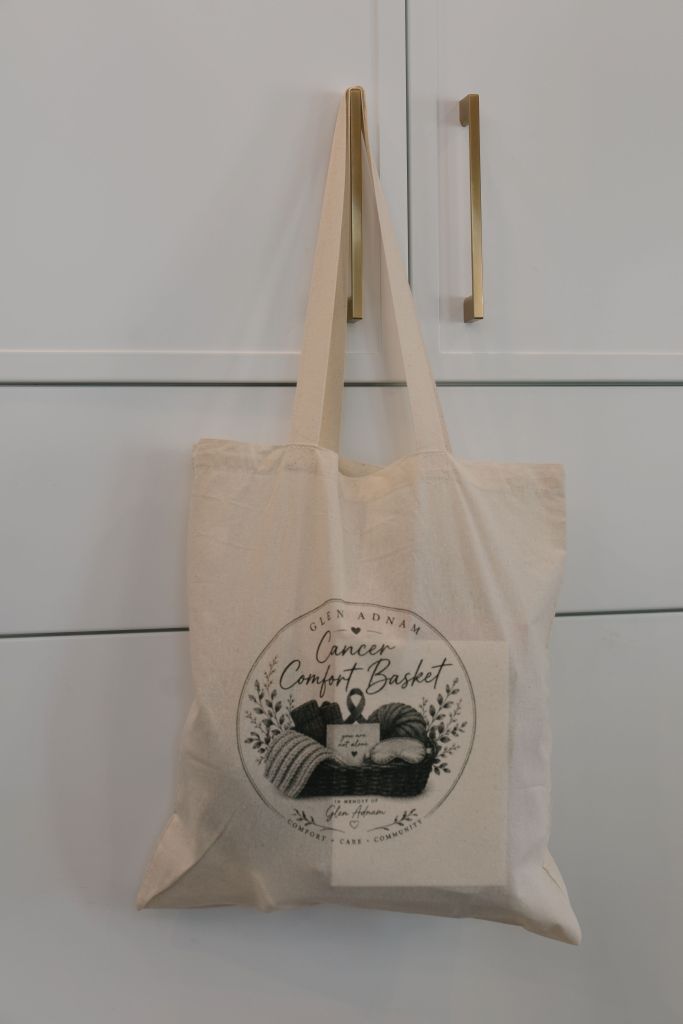 Glen Adnam Cancer Comfort Basket tote bag hanging on a cabinet, showing the branded logo with the words Comfort, Care, Community