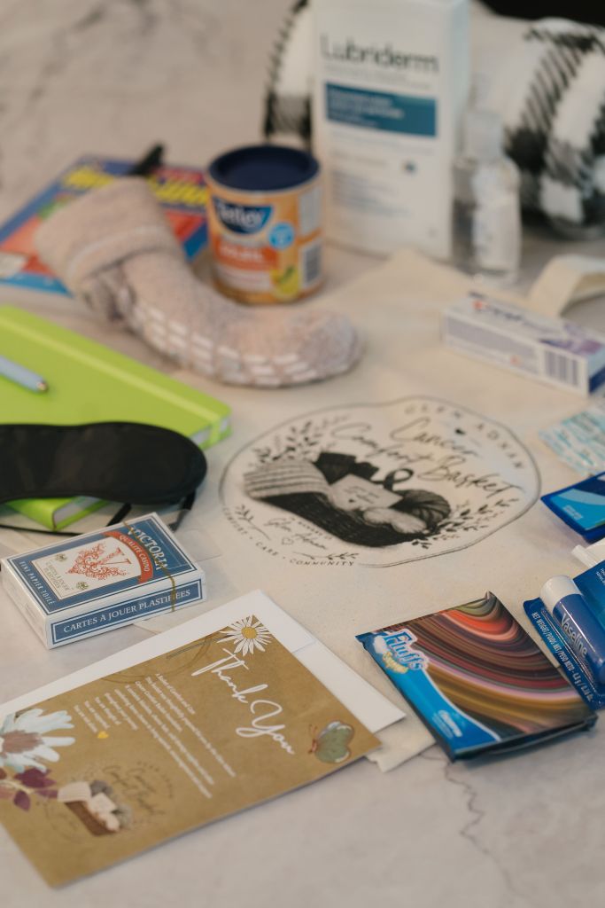 A flat lay image displaying various items including a pair of fuzzy socks, a notebook, a can of fruit, a bottle of lotion, a sleeping mask, playing cards, and a thank you card, all arranged on a marble surface.