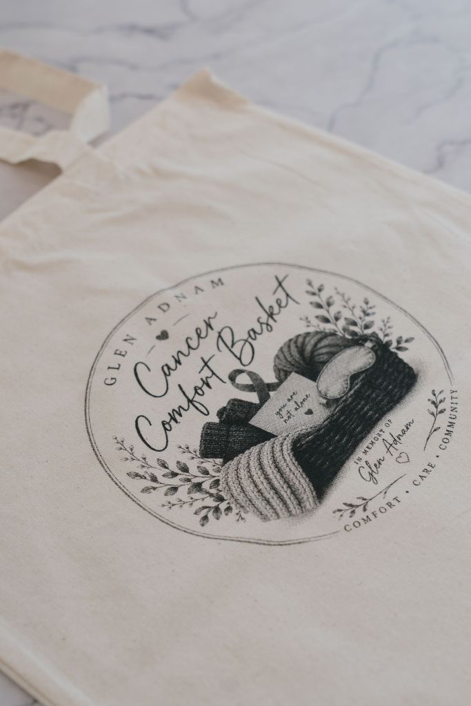Close-up of the Glen Adnam Cancer Comfort Basket tote bag laid flat, showing the hand-drawn logo with the message 'you are not alone'